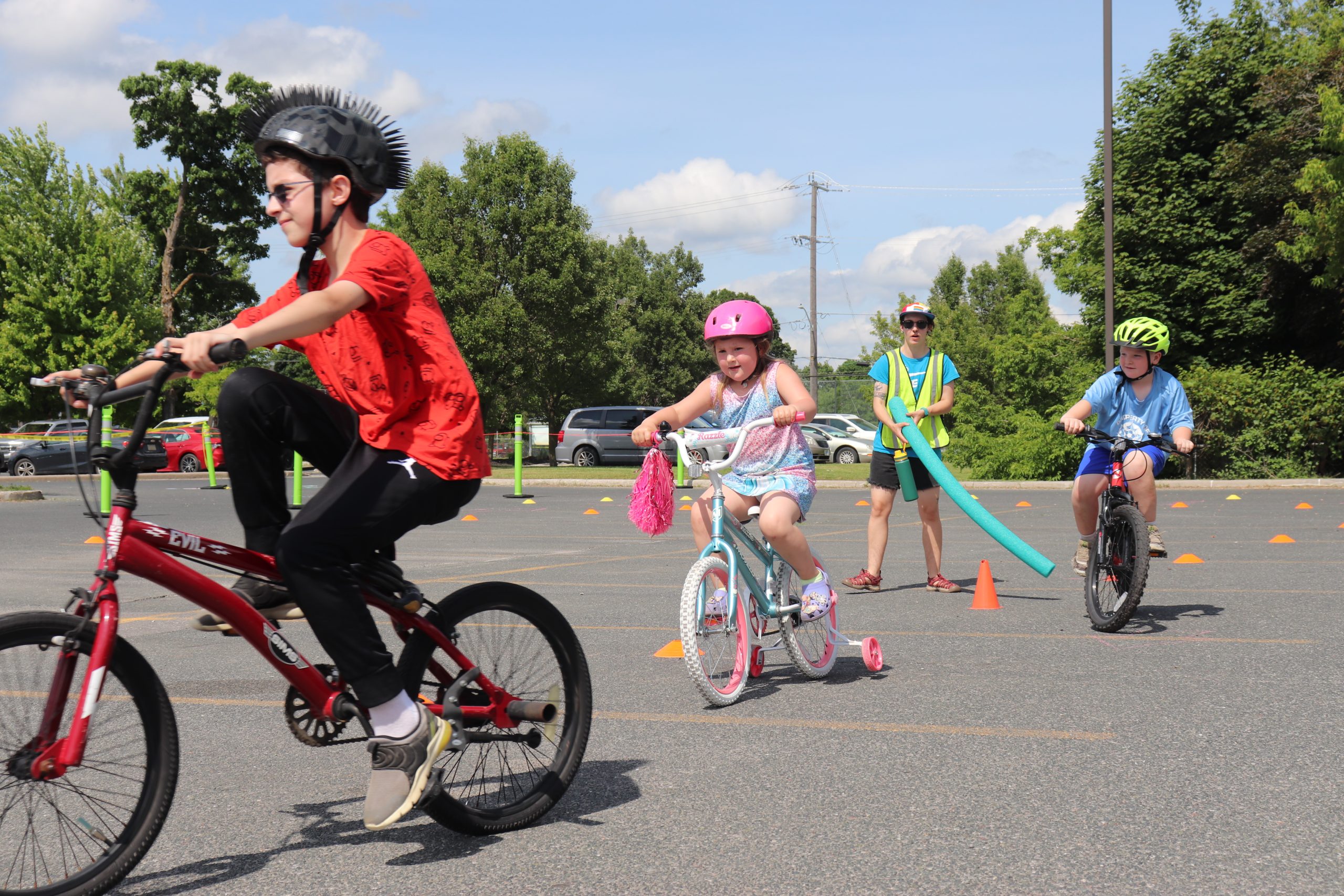 3 children are riding bikes in a parking lot on a sunny day. There are small cones set up to create a path and a young adult is standing in a green safety vest, holding a pool noodle coaching the children.