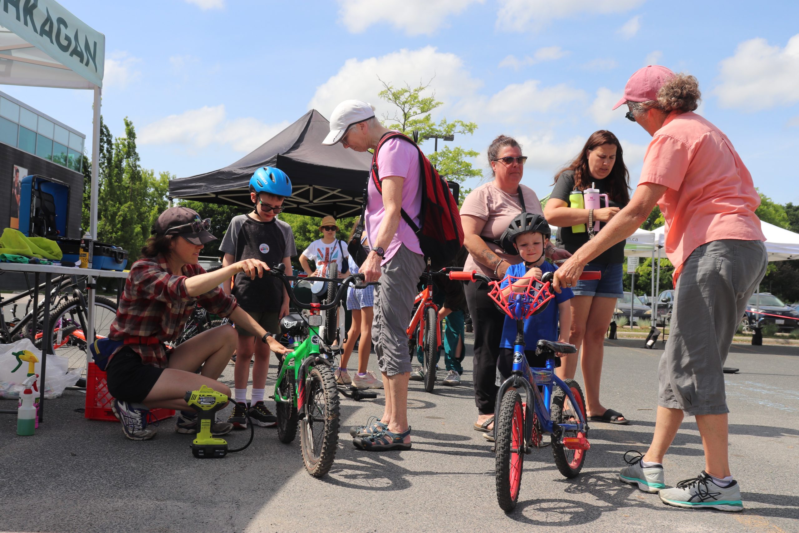 Group of people inspecting kids bikes. Everyone is dresses in functional casual clothing. From left to right: a young women in flannel and a black baseball cap, a young boy in a blue helmet, and an older person in a pink shirt and white cap. These three are standing around a green kids bike. Continuing on; a young boy in a blue shirt and black helmet, a middle aged women in a pink shirt and sunglasses, a 30-something women in a black shirt and denim shorts, and a older women in a salmon top and grey capris. These 4 are inspecting a kids spiderman bike. The sky is blue and there are a few clounds in the sky. The weather appears to be warm and everyone is dressed accordingly. This image is located in a parking lot.