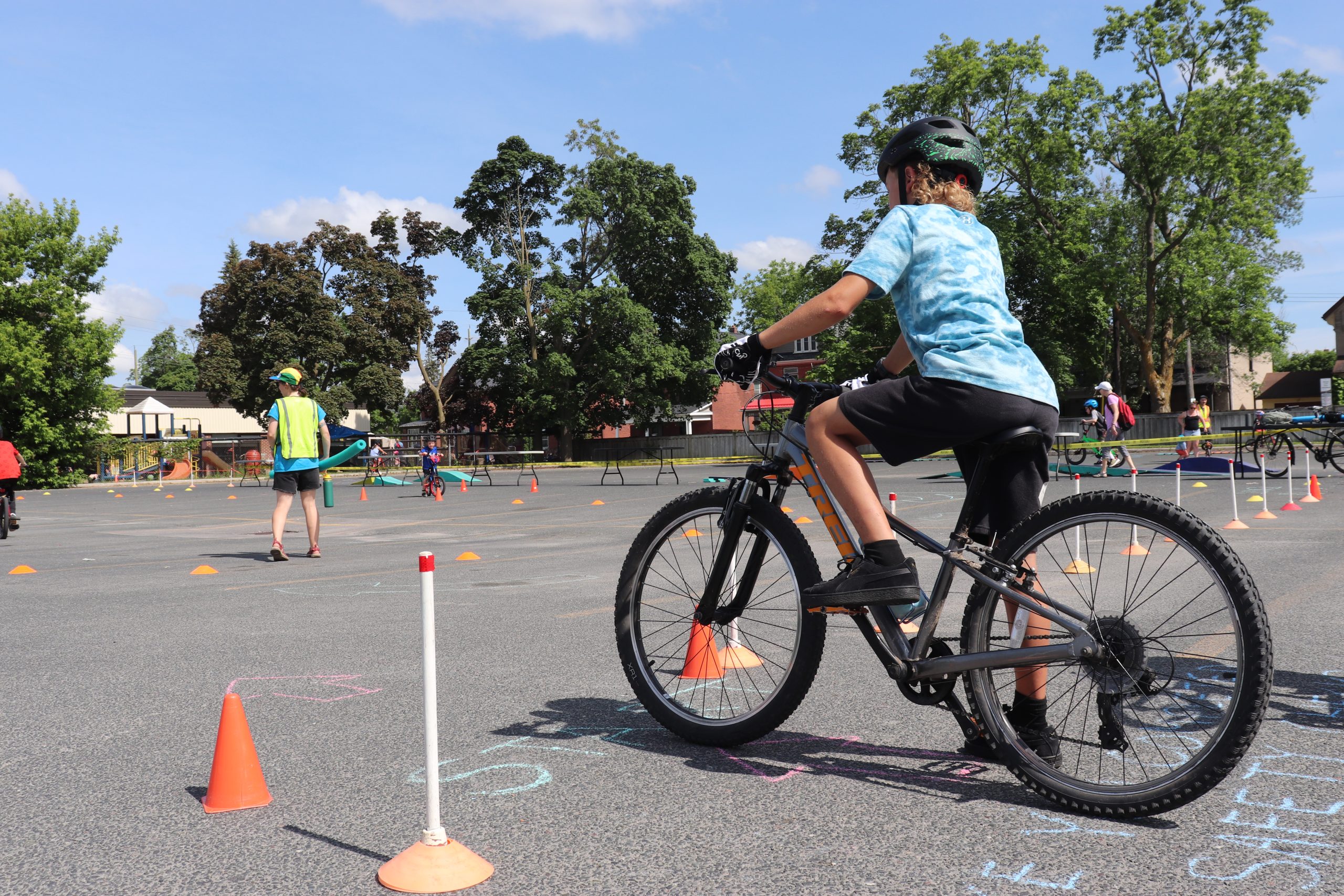 A young boy in a blue shirt and black shorts is ready to ride his black bike. There are small orange cones outlining the space in which he is to bike. In the background there are other kids on bikes and people running the event in green safety vests. The Sky is blue and the trees are full and green. the biking is happening in a parking lot with black asphalt.