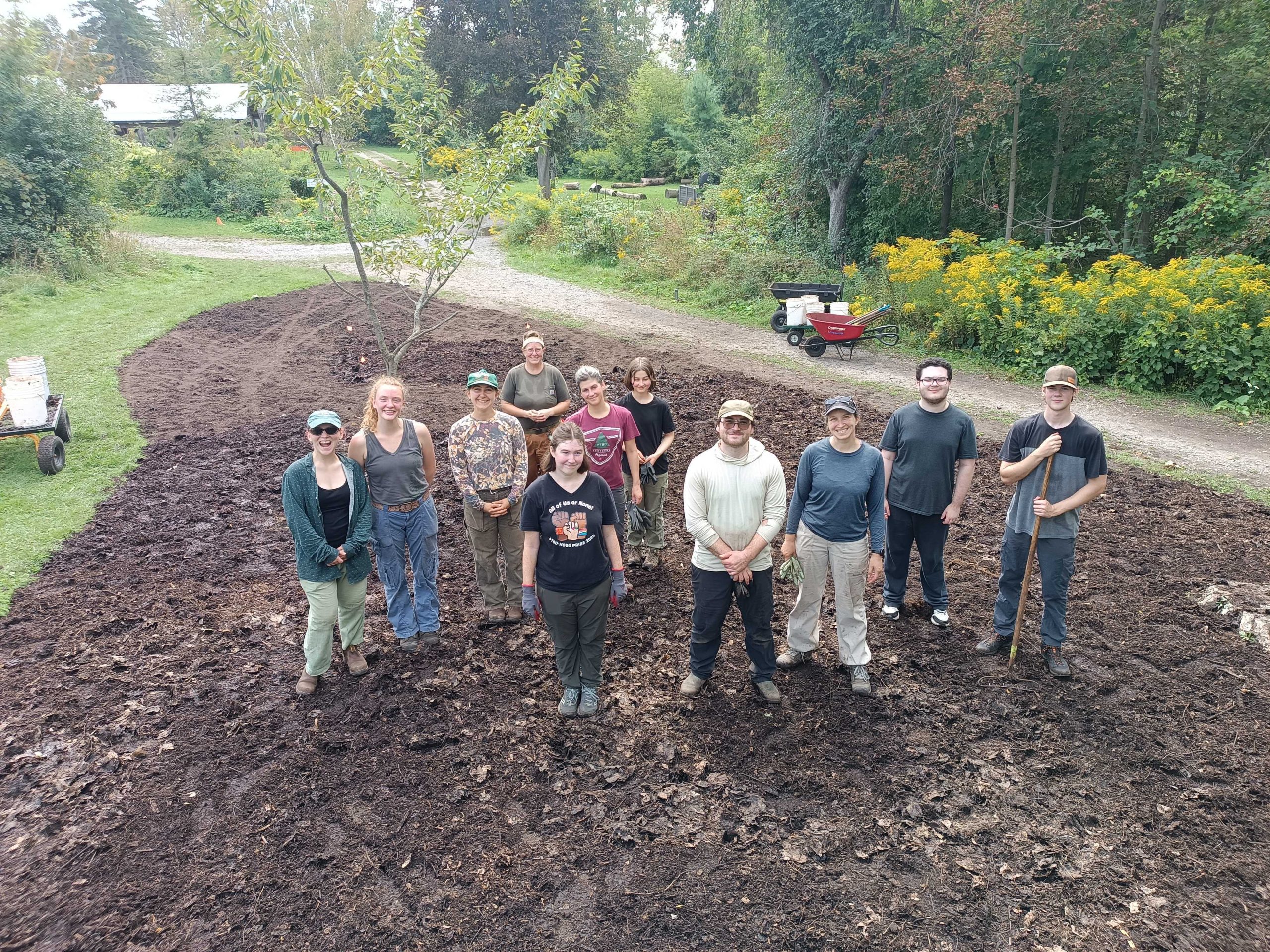 A group of ten GreenUP staff and volunteers are standing on the site of the future Little Forest at Ecology Park. Sod has been removed from the site and there is a layer of rich, dark soil and leaf mulch beneath their feet.